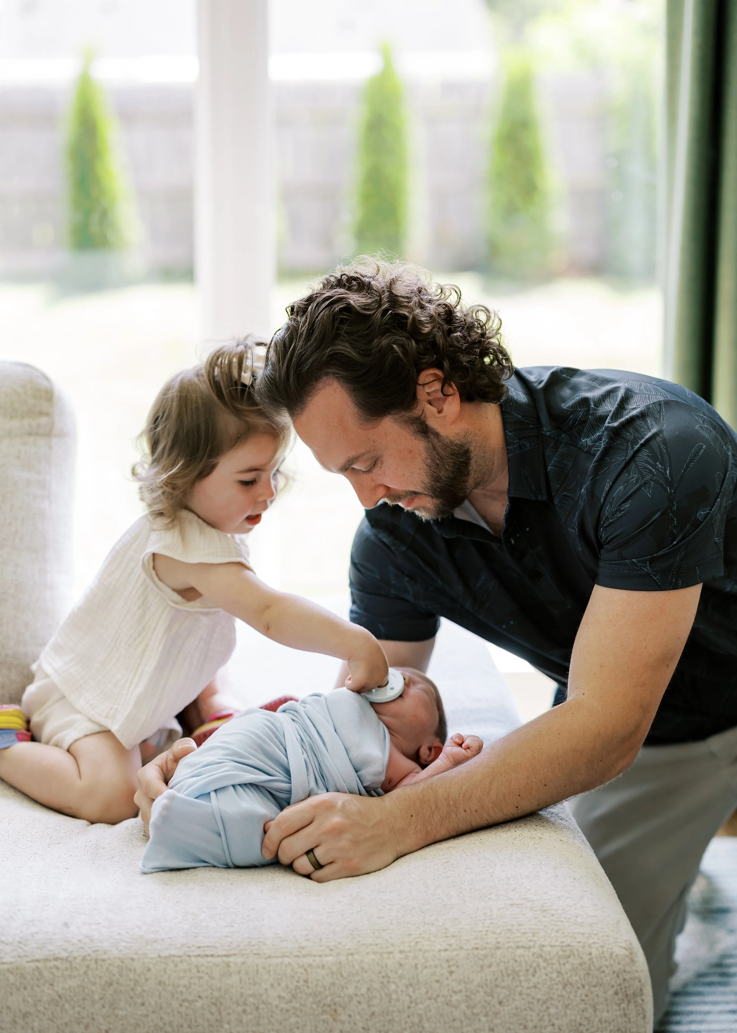 A man and two young children play with a baby on a beige couch in a bright living room with large windows and greenery outside. The man leans over and smiles at the baby, while the girl reaches for the baby and looks at him.