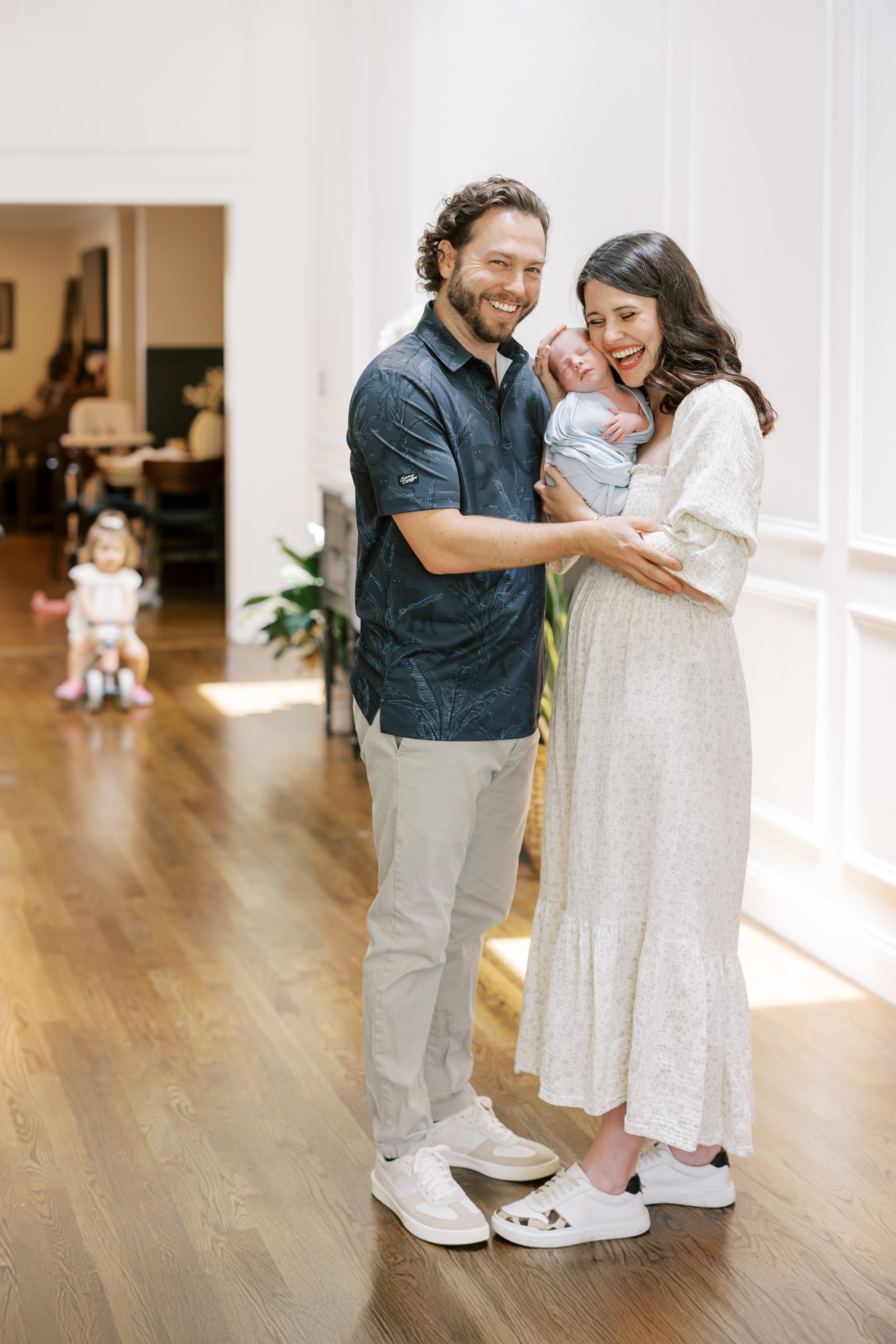 A happy couple, a man with curly hair and a beard wearing a dark Hawaiian shirt and light-colored pants, and a woman with dark curly hair in a white dress, holding their newborn baby wrapped in a light blanket, standing in a well-lit room with wooden floors and white walls.