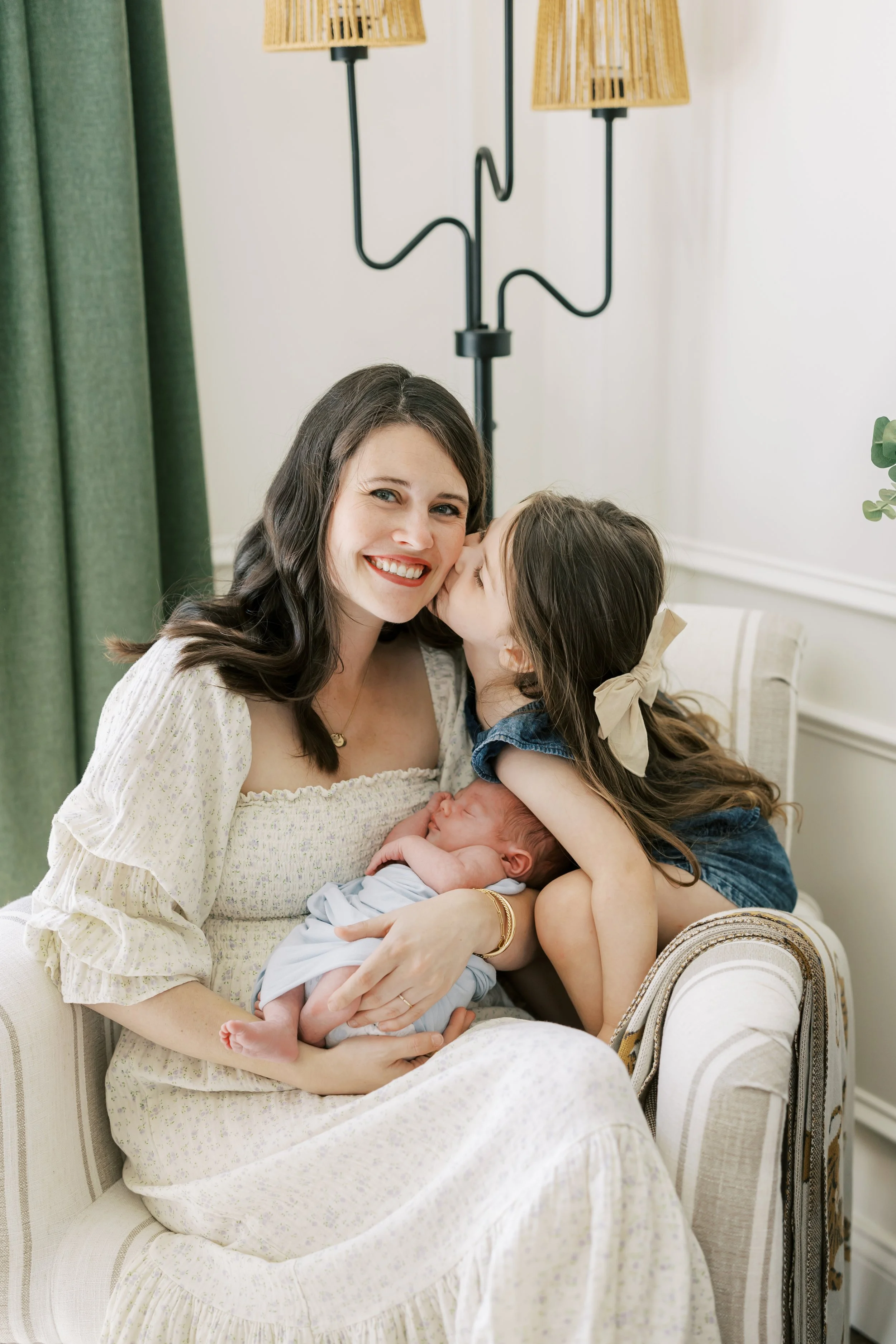 A woman with long dark hair smiling while holding a baby, with a young girl kissing her on the cheek. They are seated on an elegant cream-colored upholstered armchair. In the background, there are green curtains, wall lamps with woven shades, and a potted plant.