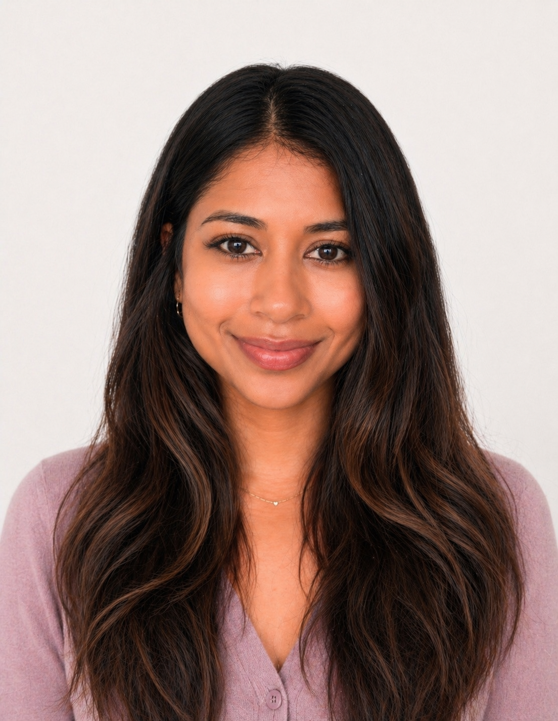 A woman with long dark wavy hair and brown eyes, wearing a pink top and a delicate necklace, smiling against a plain white background.