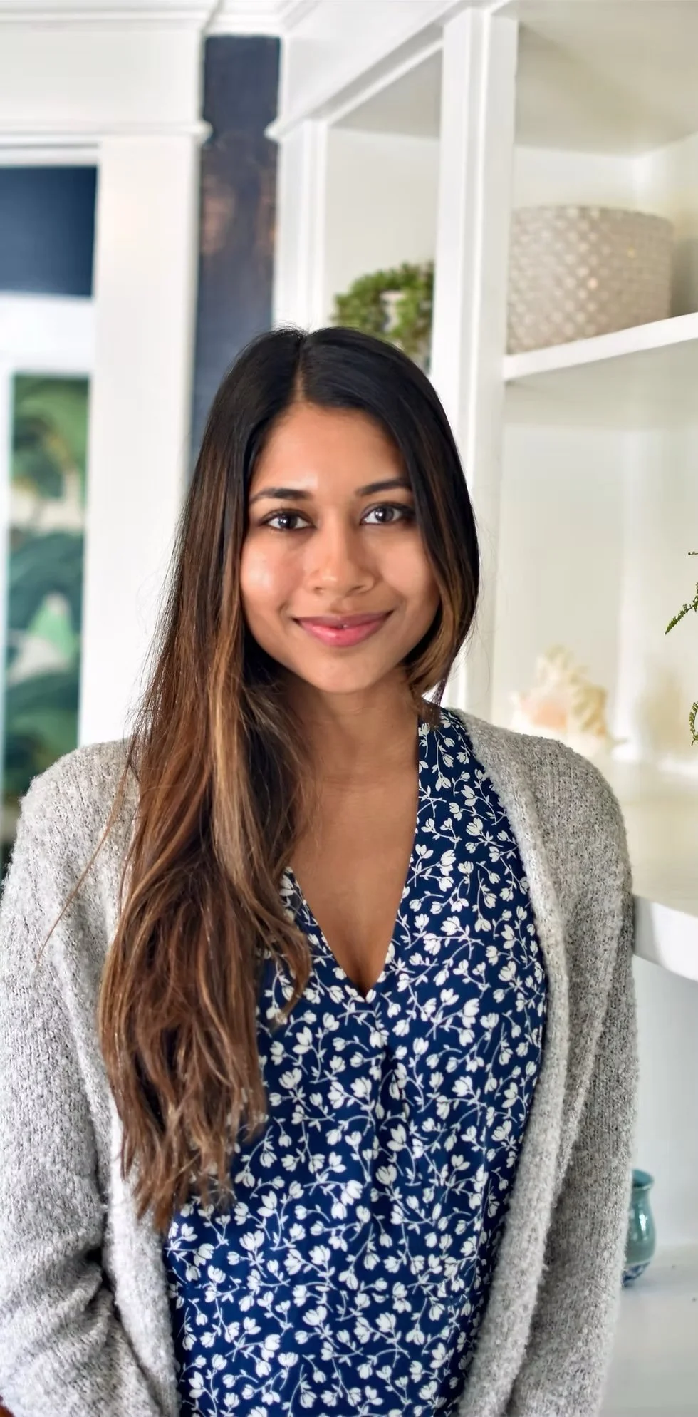 A woman with long, wavy brown hair and a warm smile, wearing a blue blouse with white floral pattern and a gray cardigan, standing indoors in a well-lit room with white shelves and green plants in the background.