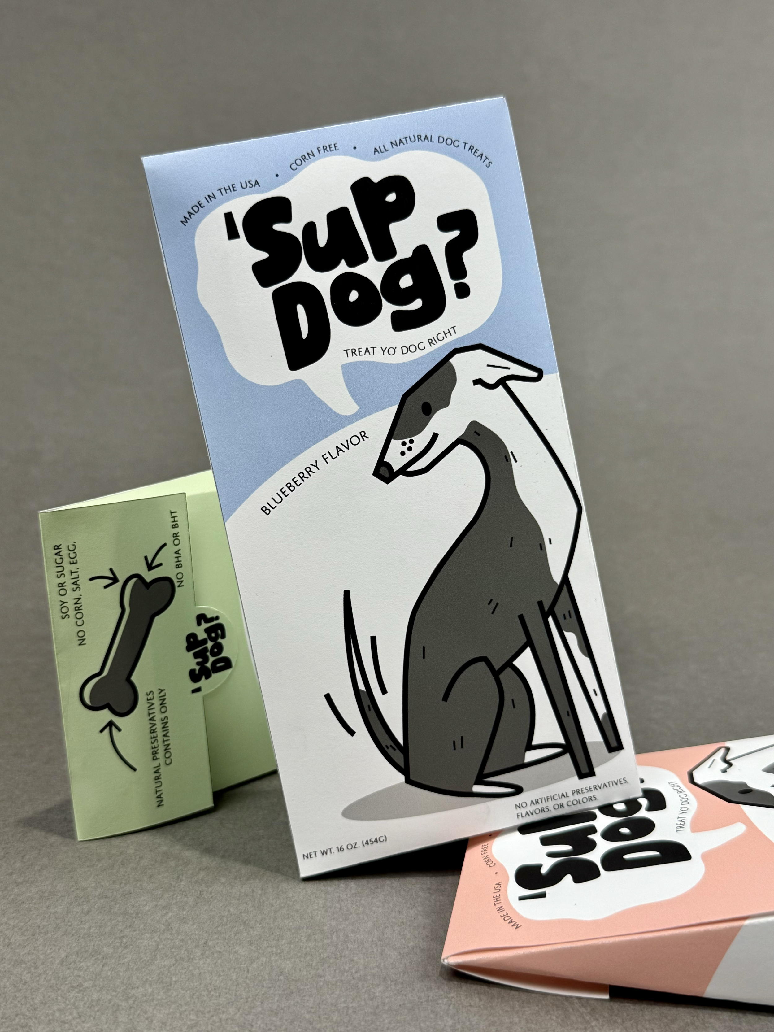 Packaging for 'Sup Dog?' blueberry flavor dog treat with a cartoon illustration of a sitting dog and a green box with a bone-shaped logo, all positioned on a gray surface with a plain background.