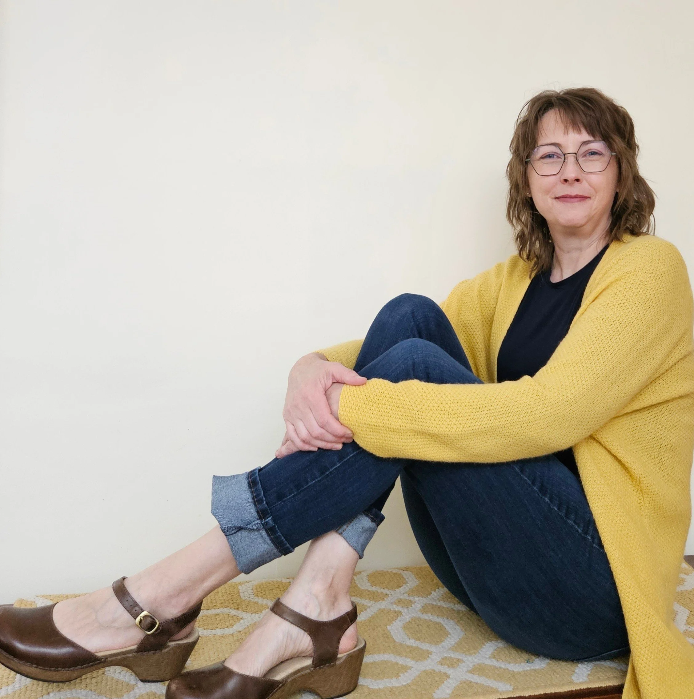 A woman with curly brown hair and glasses is sitting on the floor against a yellow blanket or rug, smiling. She is wearing a yellow cardigan over a black top. Two other people's legs and hands are visible, one with a dark skirt and brown sandals and the other barefoot, reaching to hold her hand. The floor has a beige patterned rug and the background is a plain light-colored wall.