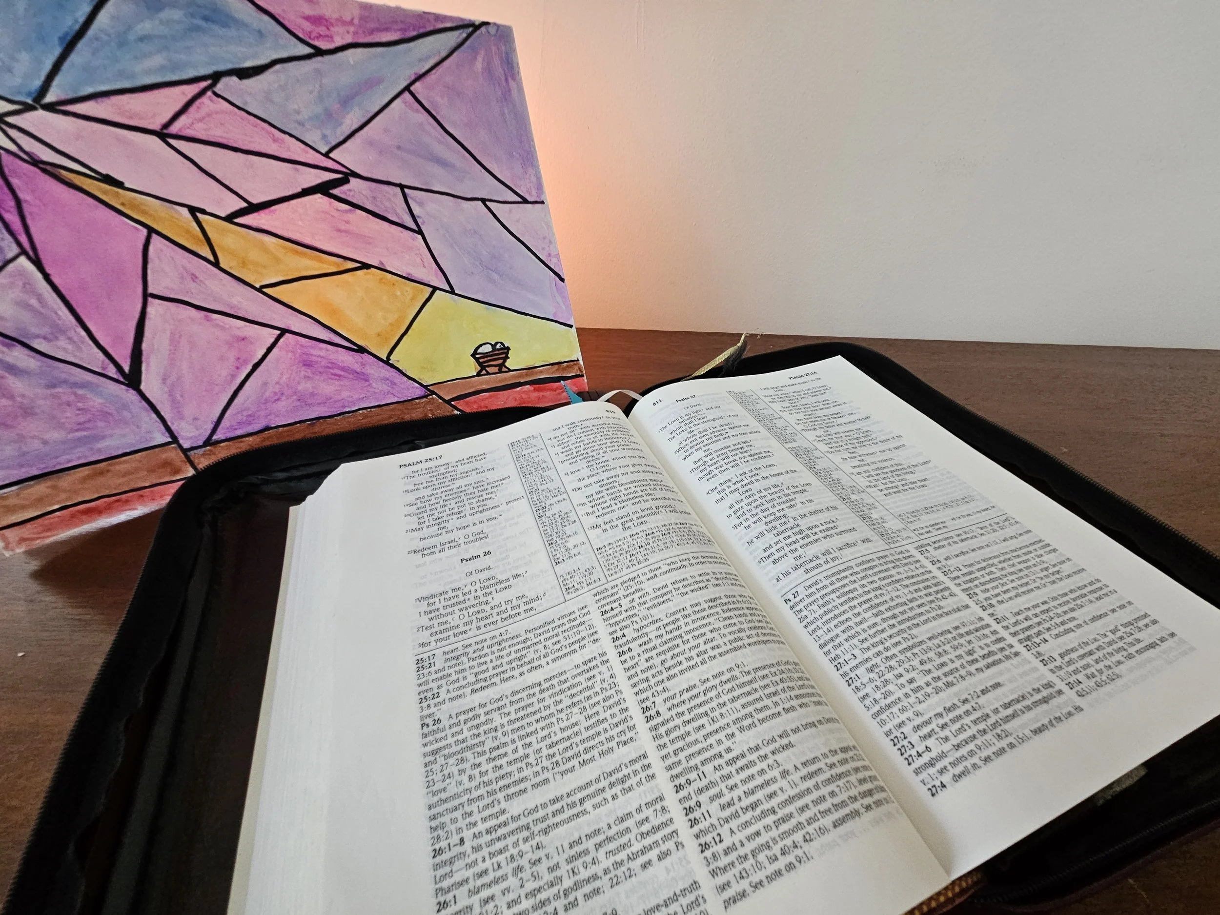 Open Bible on a wooden table with a colorful stained glass artwork in the background, featuring geometric shapes in purple, pink, yellow, and orange.