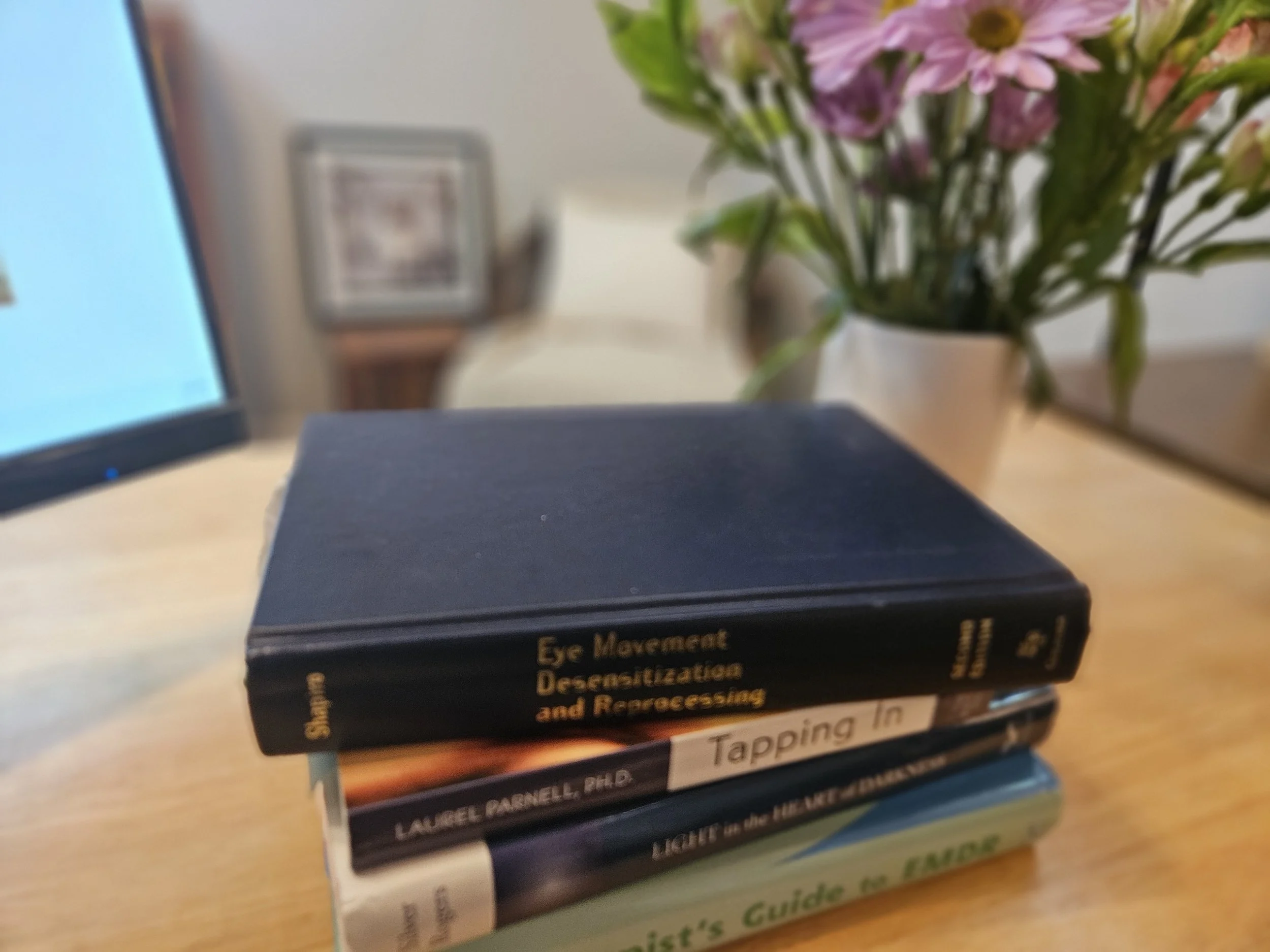 Stack of books on a wooden surface, with a blurred background of a flower vase and framed picture. The visible book titles include 'Eye Movement Desensitization and Reprocessing' and others.