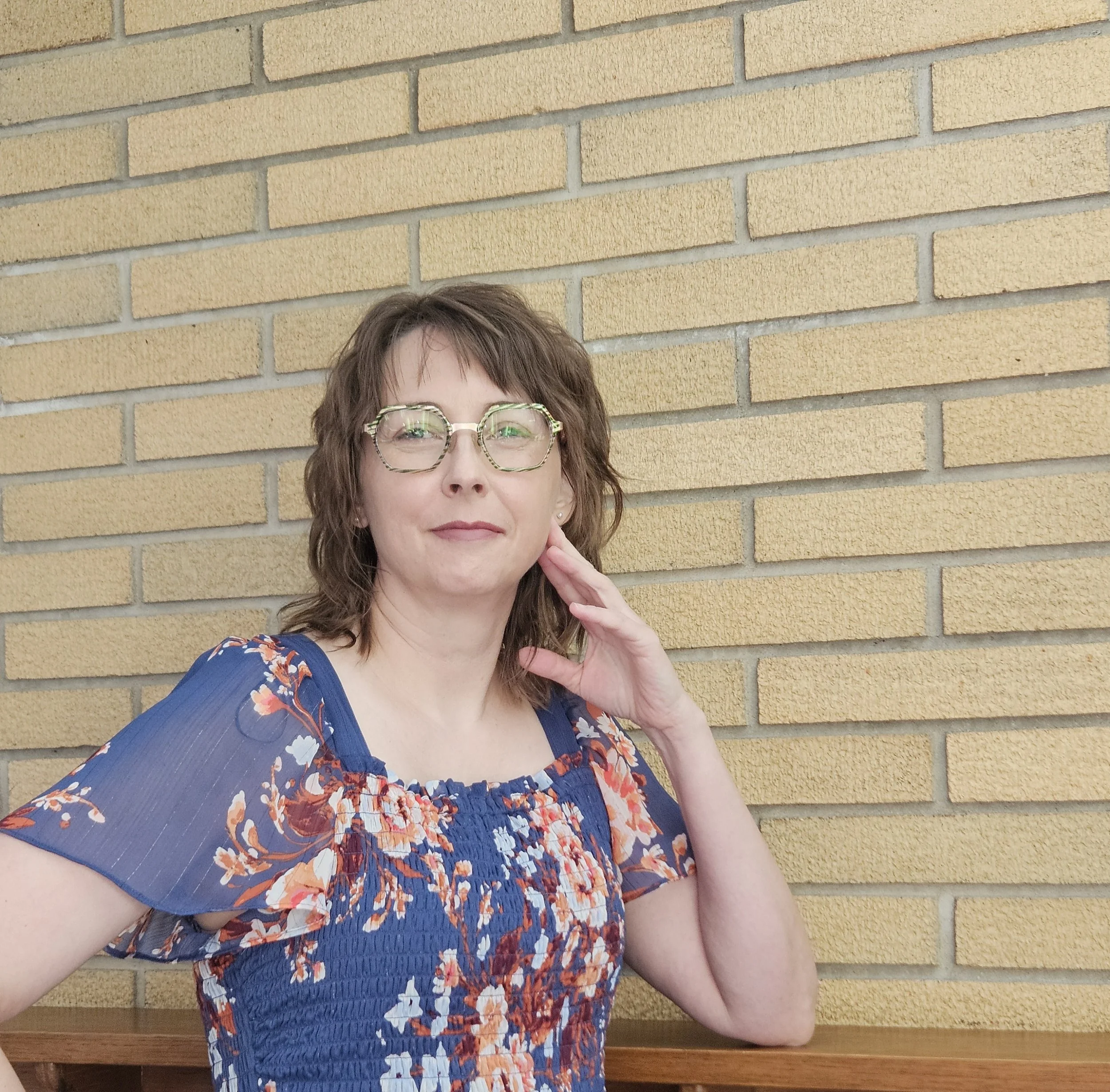 Woman with glasses and short brown hair, wearing a blue floral dress, standing in front of a yellow brick wall.