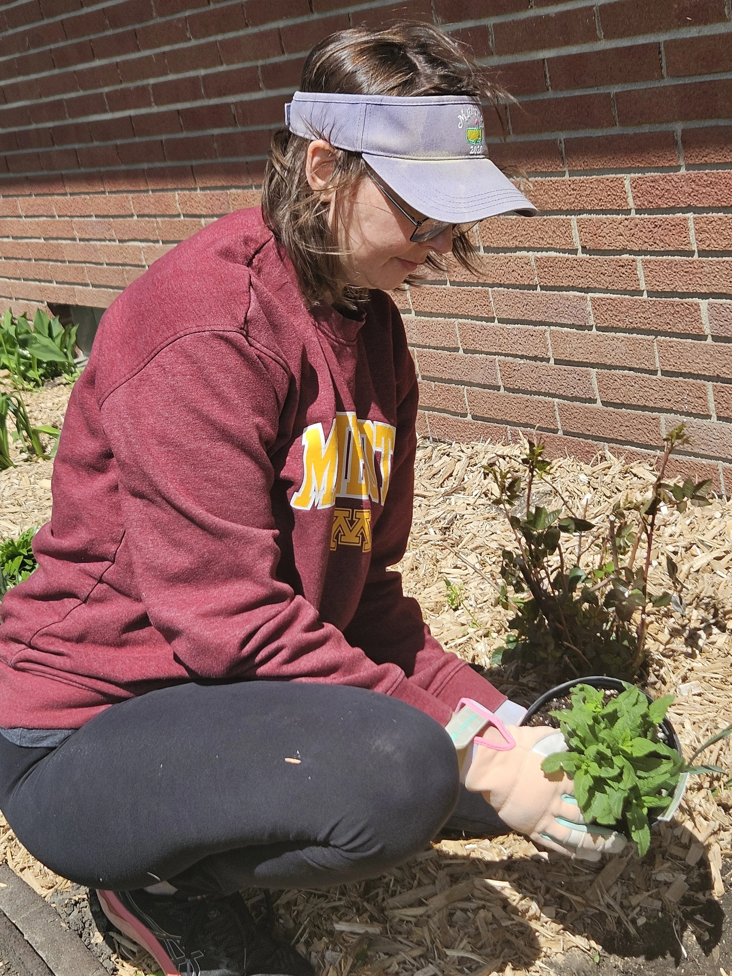 A person wearing a purple visor and glasses, dressed in a maroon sweatshirt and black pants, kneeling outside near a red brick wall and gardening plants.