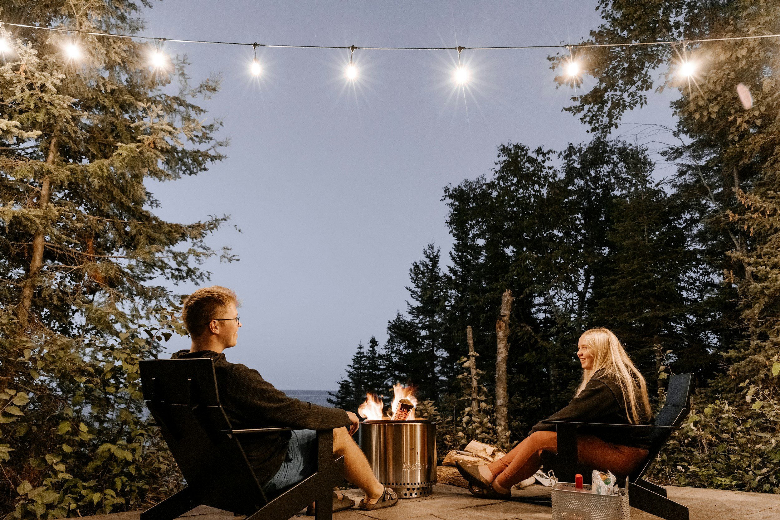 Two people sitting outdoors around a small fire at night, surrounded by trees and string lights overhead. The man on the left is smiling, and the woman on the right is smiling back. They are seated in chairs with a table nearby holding some items.