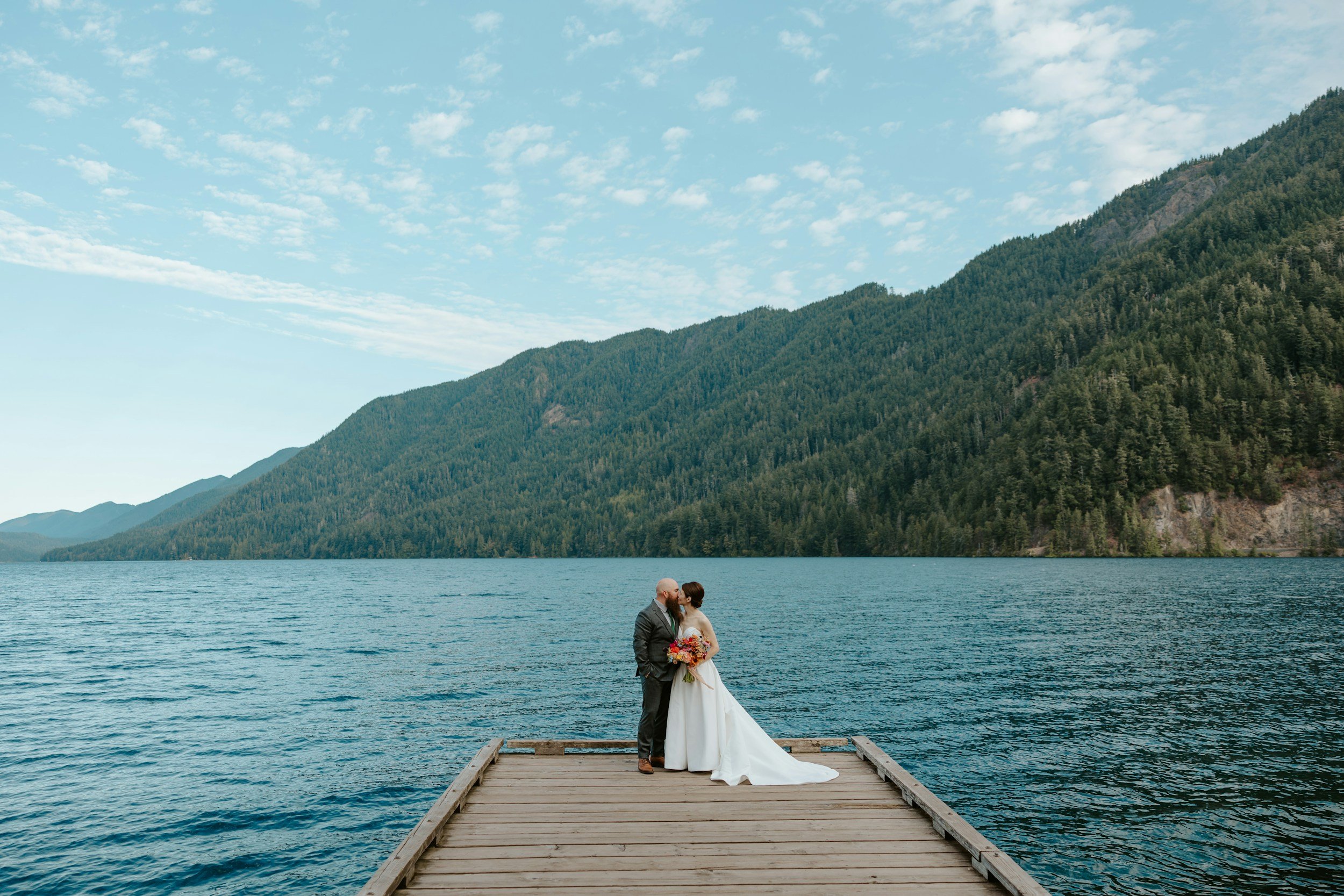 A couple dressed in wedding attire, standing on a wooden dock by a large lake, with a backdrop of green mountains and a partly cloudy sky.