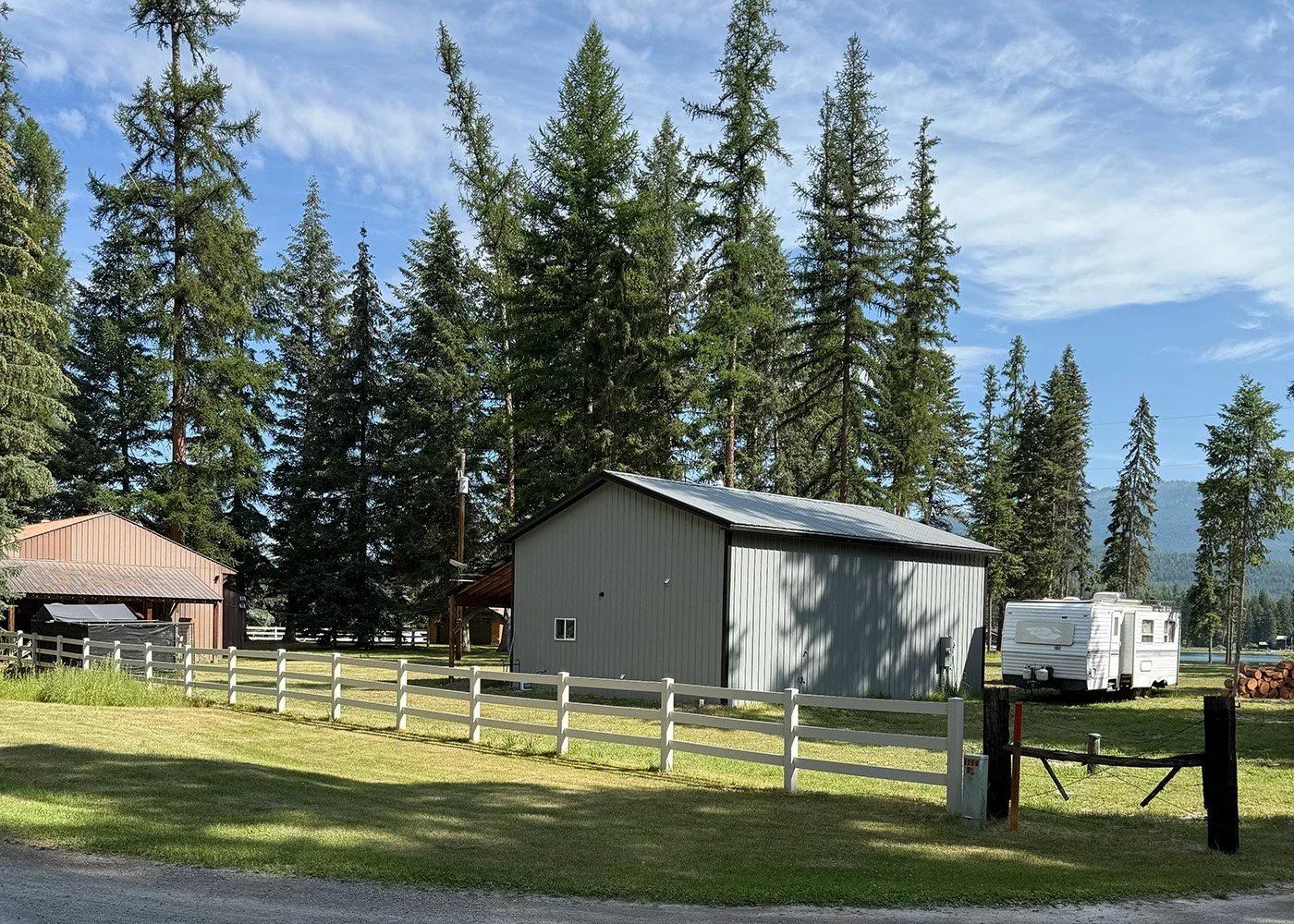 A rural scene with a large metal shed, a white camper trailer, and a fenced grassy yard surrounded by tall pine trees under a partly cloudy sky.