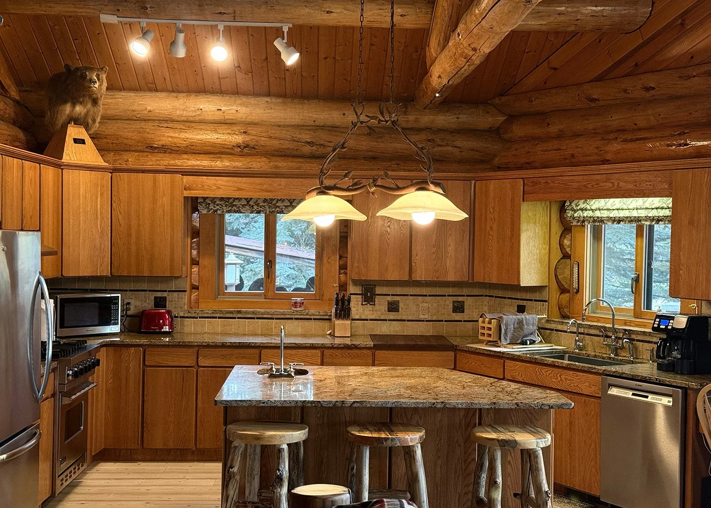 Cozy rustic kitchen with wooden cabinetry and log ceiling, featuring a central island with bar stools, stainless steel appliances, and windows with view of trees outside.