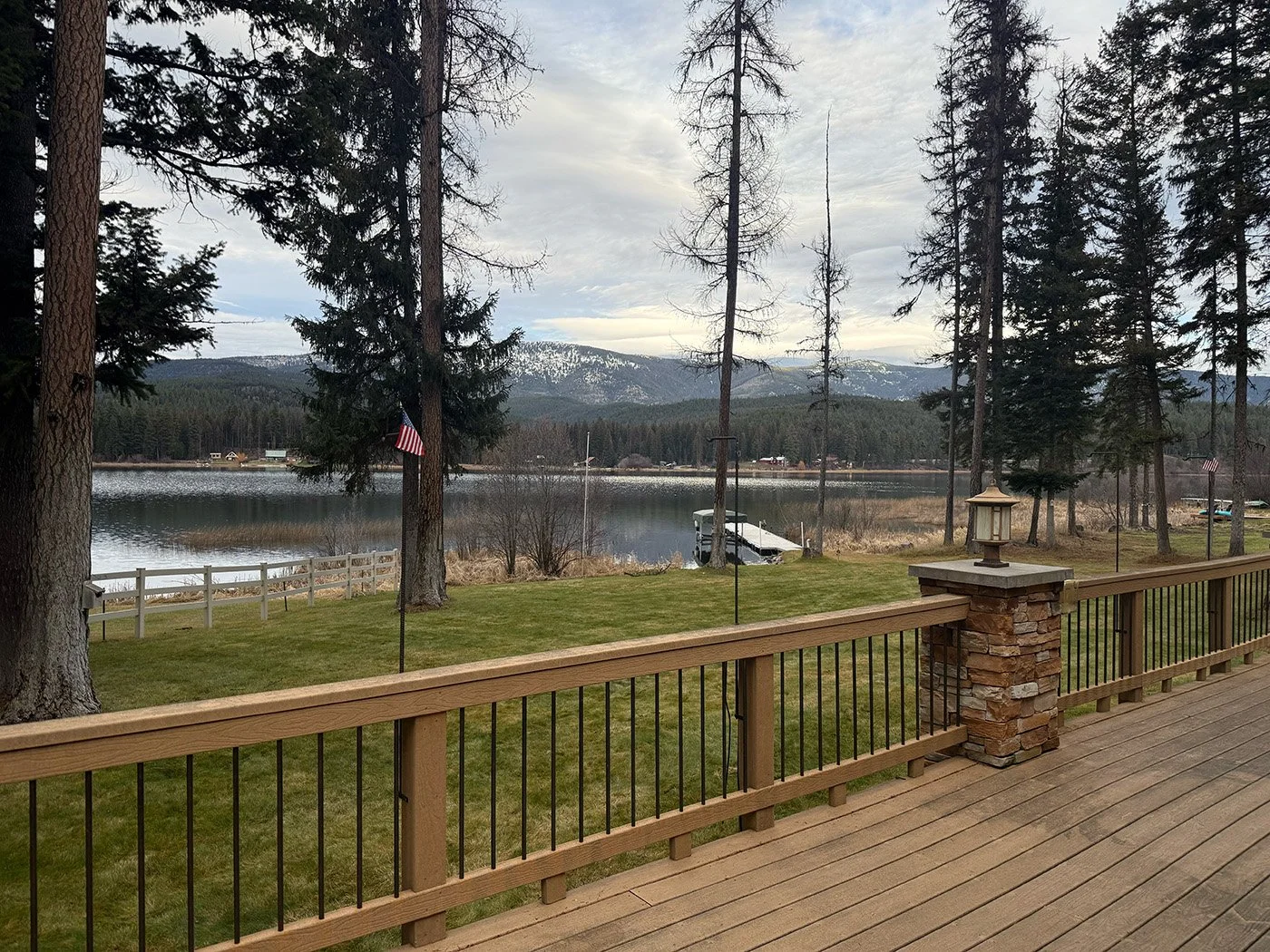 View of a lake with mountains in the background, trees surrounding the shoreline, a dock, American flags, a grassy area, and part of a wooden deck with a lantern on a stone pillar.