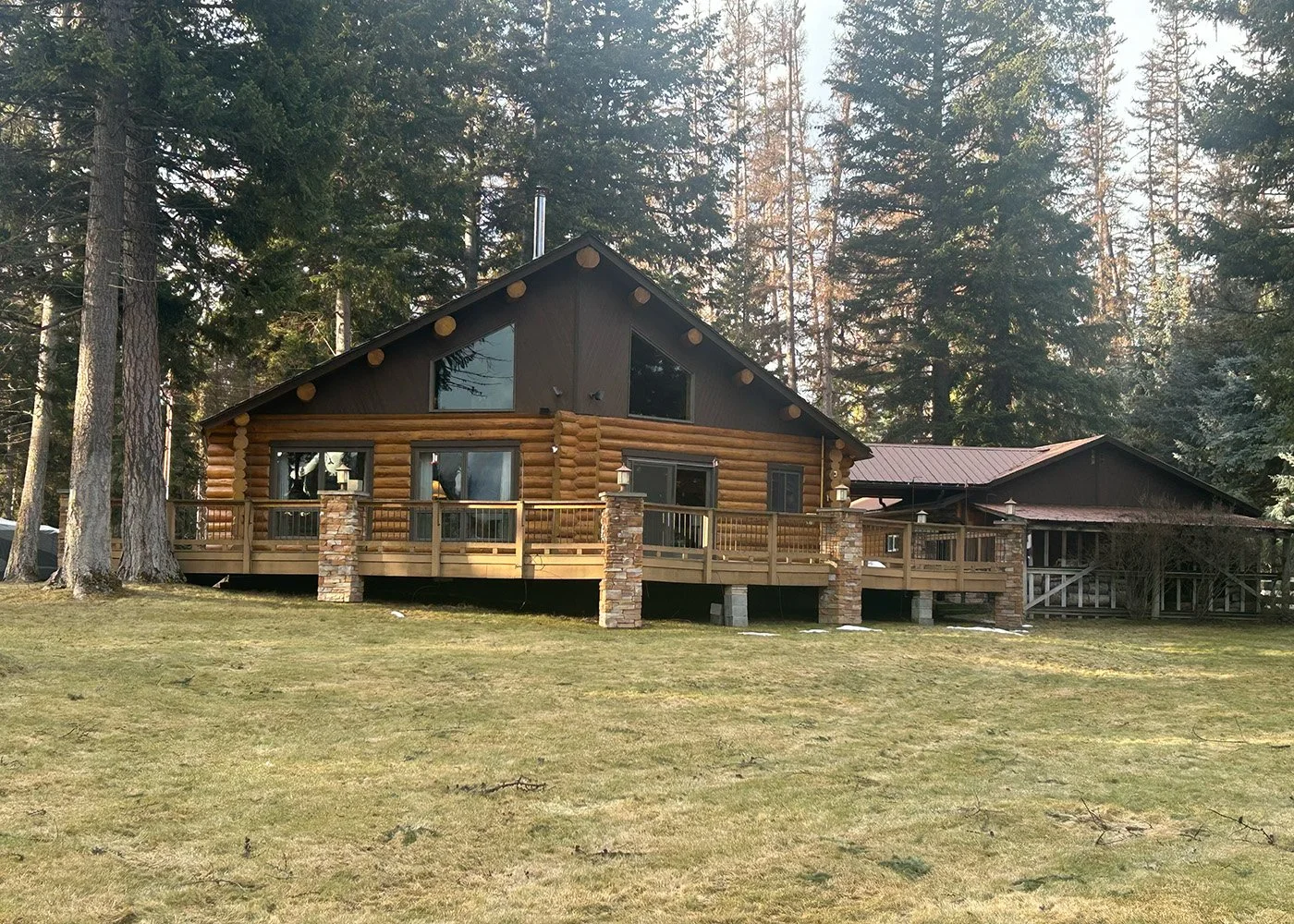 A log cabin-style house with a wooden deck and stone pillars, set among tall trees on a grassy lawn.
