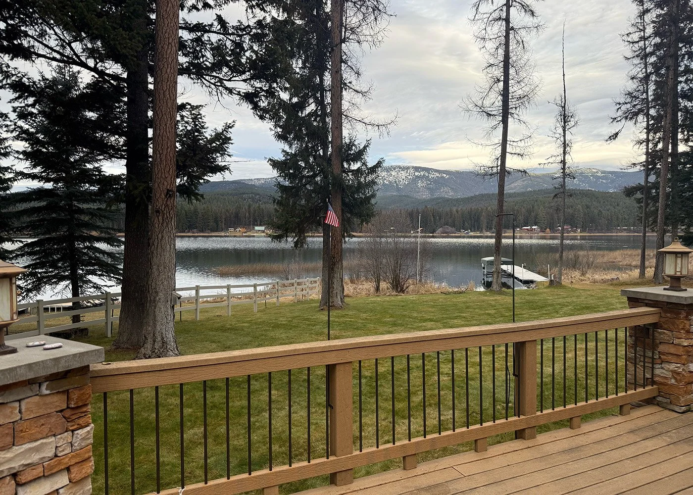 A backyard view overlooking a lake with trees and snow-capped mountains in the background, taken from a wooden deck with a railing.