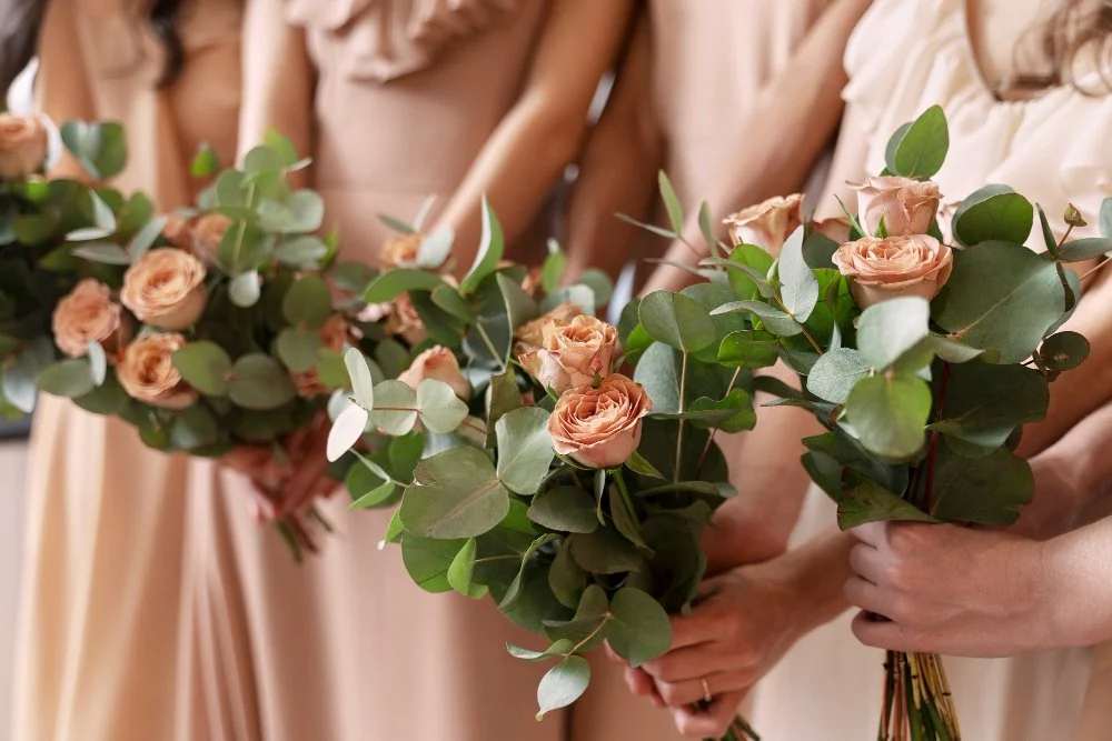 Women in beige dresses holding bouquets of pink roses and eucalyptus leaves.