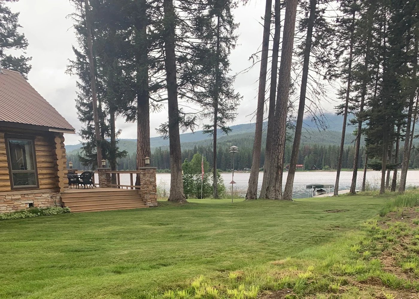 A lakeside yard with a wooden house on the left, a deck with outdoor furniture, tall pine trees, a lake, a dock, and mountains in the background.