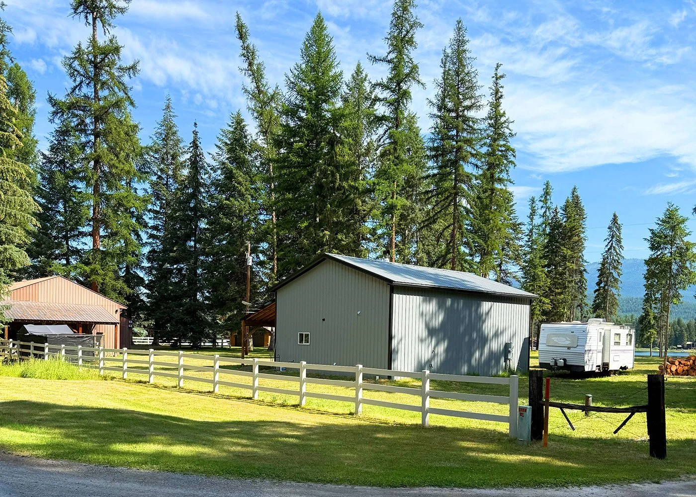 A rural scene with a grassy yard, white fence, two buildings, a camper trailer, and tall pine trees under a partly cloudy sky.