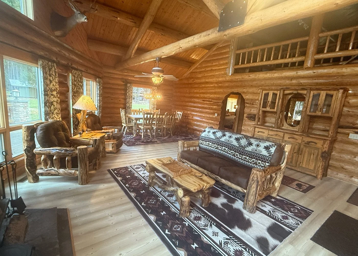 Interior of a rustic log cabin living room with log furniture, cowhide rugs, a patterned blanket on a sofa, and a dining area in the background, all with wood paneling and large windows.