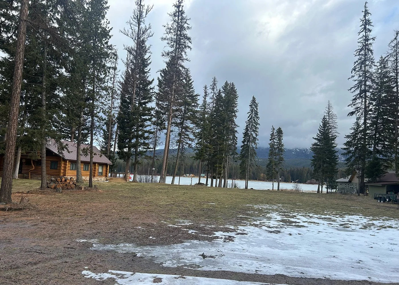 A peaceful lakeside scene with a wooden cabin, tall pine trees, and a partly frozen lake in the background.