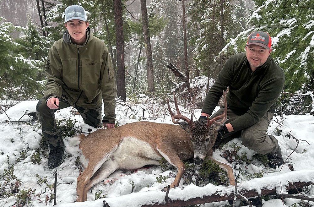 Two men in winter clothing kneel on snow-covered ground next to a freshly killed deer with antlers in a snowy forest.