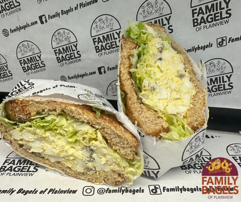 Close-up of a lettuce and egg salad sandwich in a toasted bun, wrapped in branded paper with Family Bagels of Plainview logo, on a paper with the same branding.