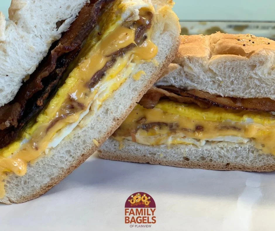 Close-up of a breakfast bagel sandwich with eggs, bacon, and cheese, cut in half to show the filling. The bagel is toasted and sesame seed-topped. The logo 'Family Bagels of Plainview' is visible at the bottom of the image.