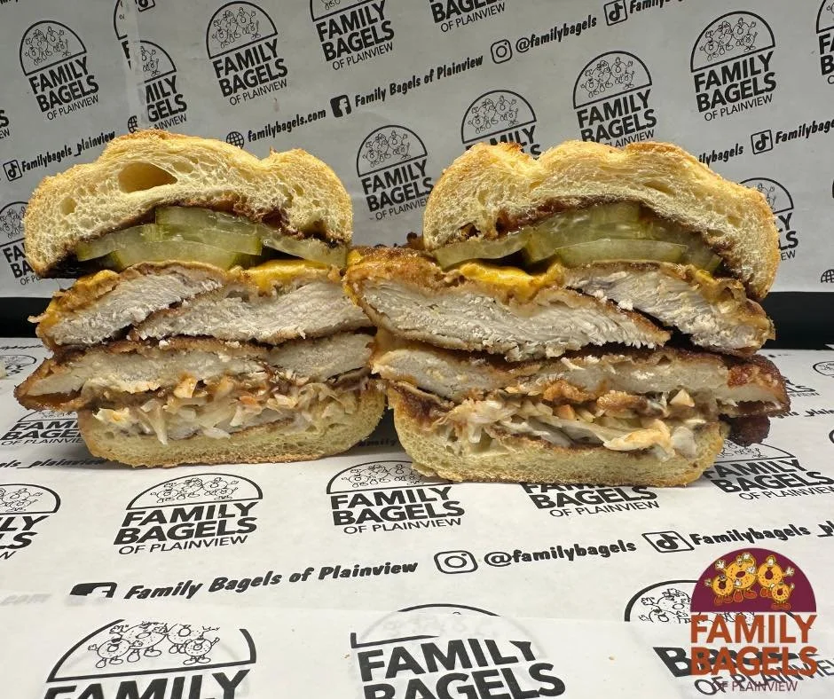 Half of a large sandwich with breaded chicken fillet, pickles, and melted cheese inside a sandwich bun, placed on branded paper with bakery logo in the background.