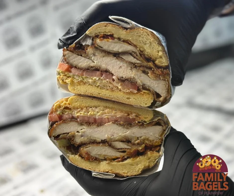Cross-section of a double-decker sandwich with fried chicken, bacon, and egg on a bagel, held by a person wearing black gloves. The bagel is from Family Bagels of Plainview.