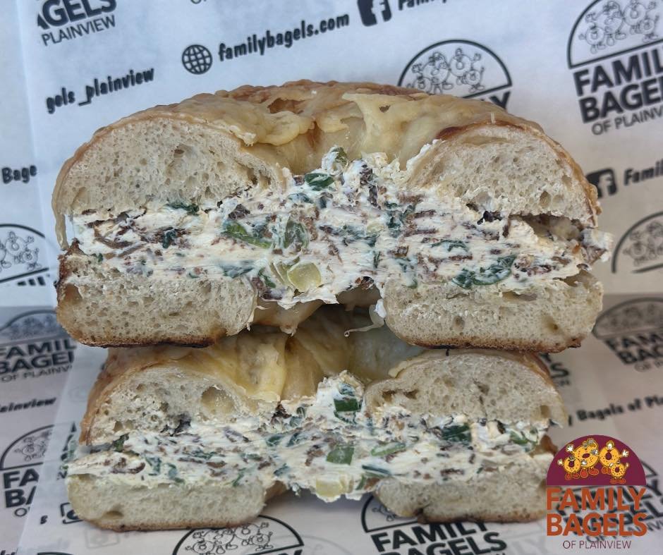 A bagel sandwich with cream cheese, green onions, and chopped sun-dried tomatoes, cut in half and stacked on a branded paper from Family Bagels of Plainview.