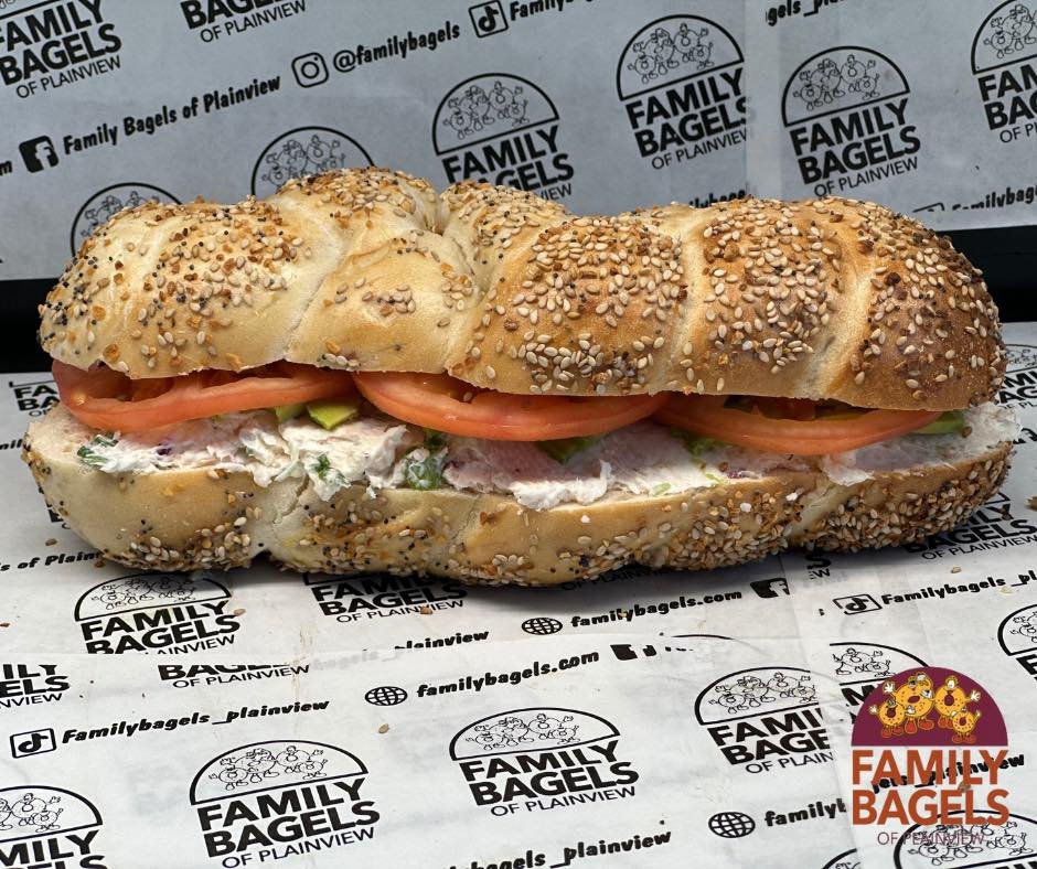 A salted sesame bagel sandwich with sliced tomatoes, lettuce, and chicken salad inside, placed on branded paper from Family Bagels.