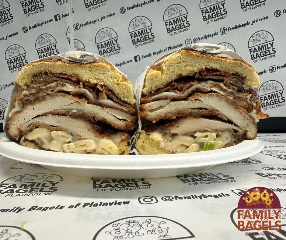 Cross-section of a large, layered sandwich with meats, cheeses, and vegetables, served on a white foam plate, with a branded background and logo of Family Bagels of Plainview.