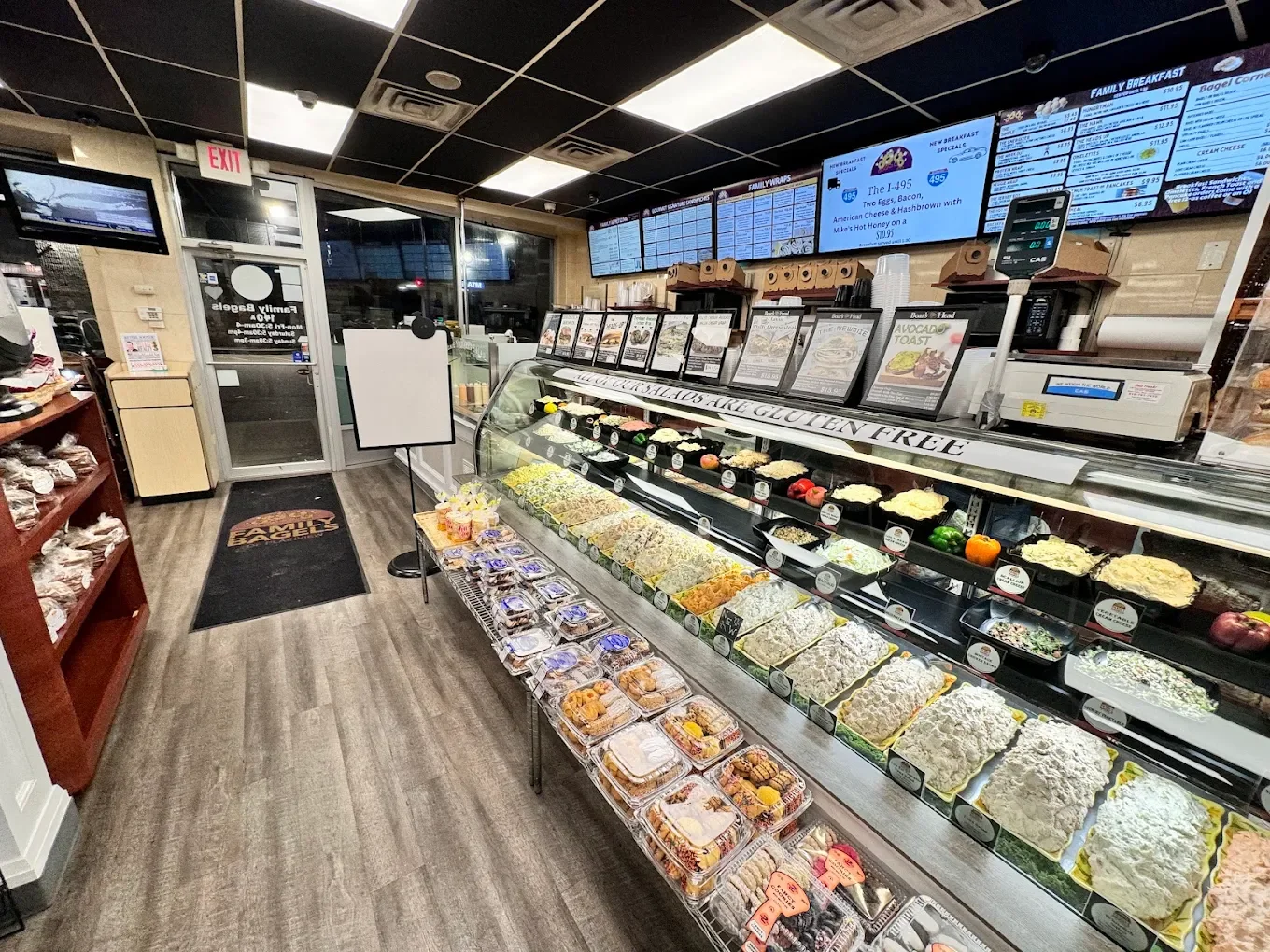 Interior of a deli or sandwich shop with a glass display case filled with salads and deli items, menu boards on the wall, and baked goods on a table near the door.