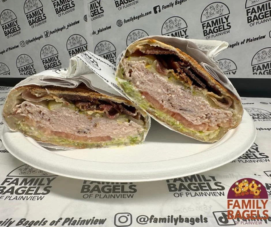 Cross-section of a bagel sandwich with turkey, lettuce, tomatoes, onions, and cheese on a white plate. Background has branding for Family Bagels of Plainview.