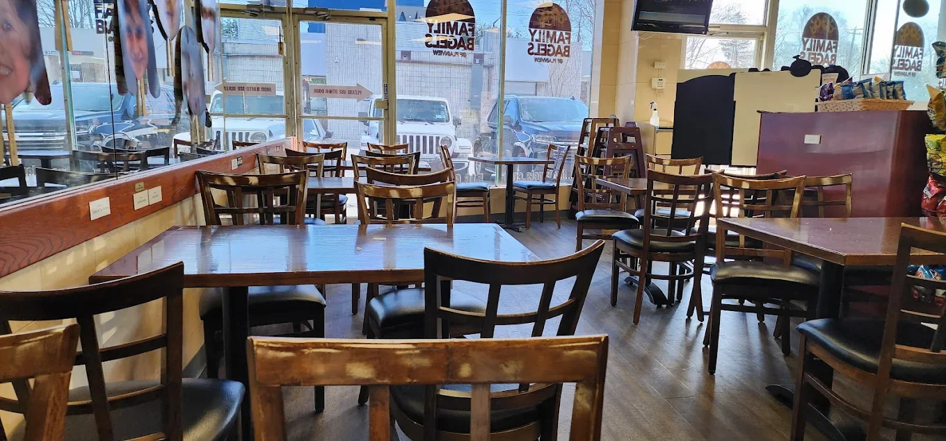 An empty restaurant with wooden tables and chairs, large glass windows, and sunlight coming through. Decor includes hanging picture cutouts and signs on the window.