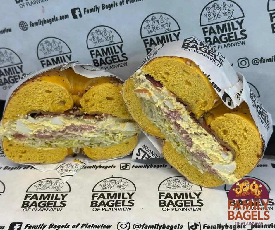 A sliced sandwich on yellow bread with lettuce, meats, and mayonnaise inside, wrapped in branded paper from Family Bagels, placed on branded paper wrap from Family Bagels.