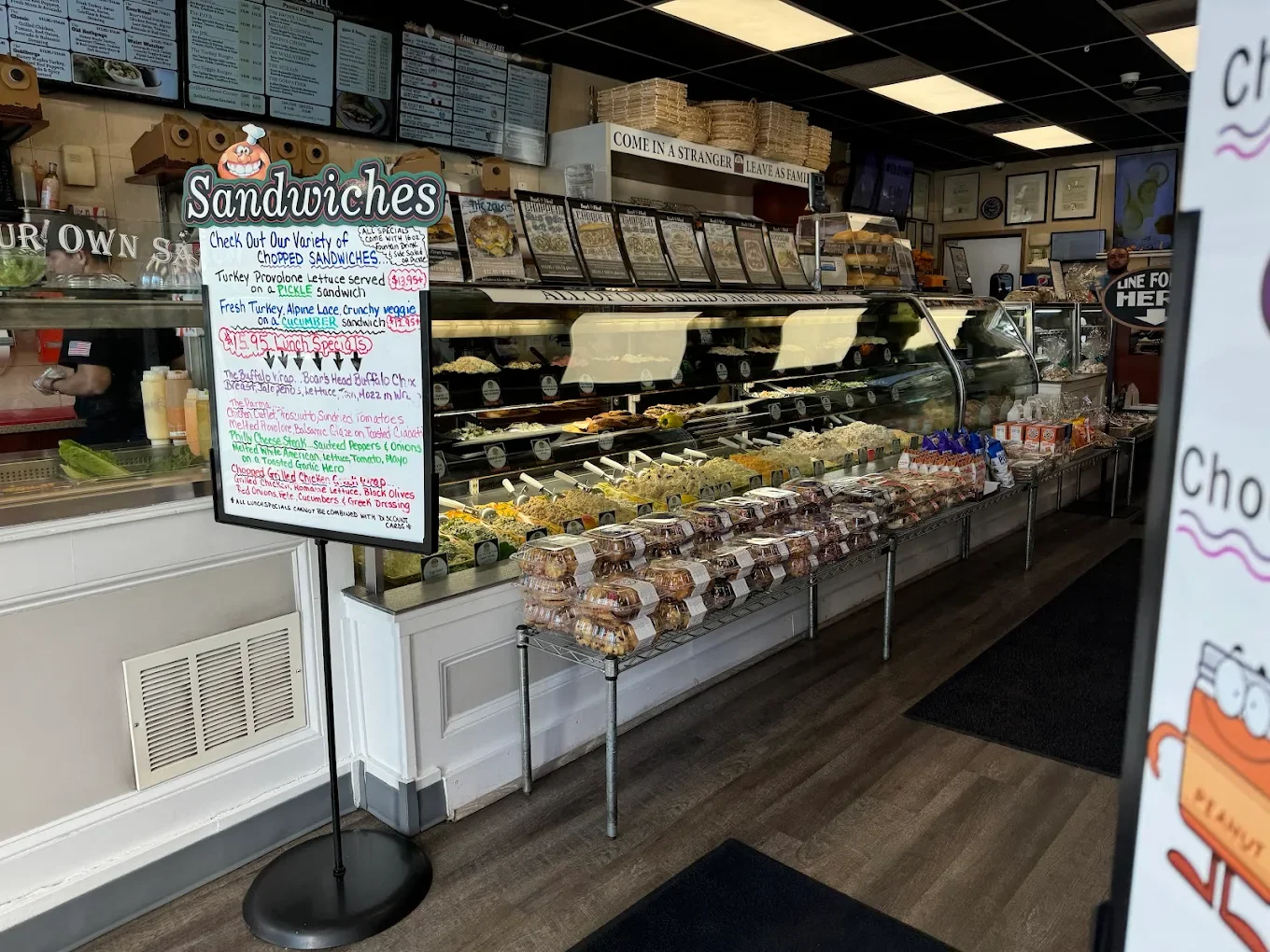 Inside a sandwich shop with a display case filled with various sandwiches, a menu on the wall, and a signboard listing sandwich options and lunch specials.