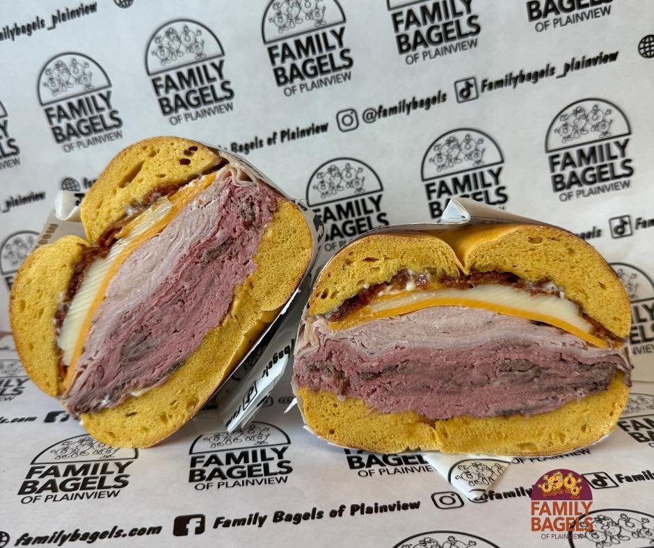 Cross section of a bagel sandwich with layers of smoked meat, cheese, and possibly egg, inside a toasted bagel with a sesame seed topping, on branded paper with 'Family Bagels of Plainview' logo.
