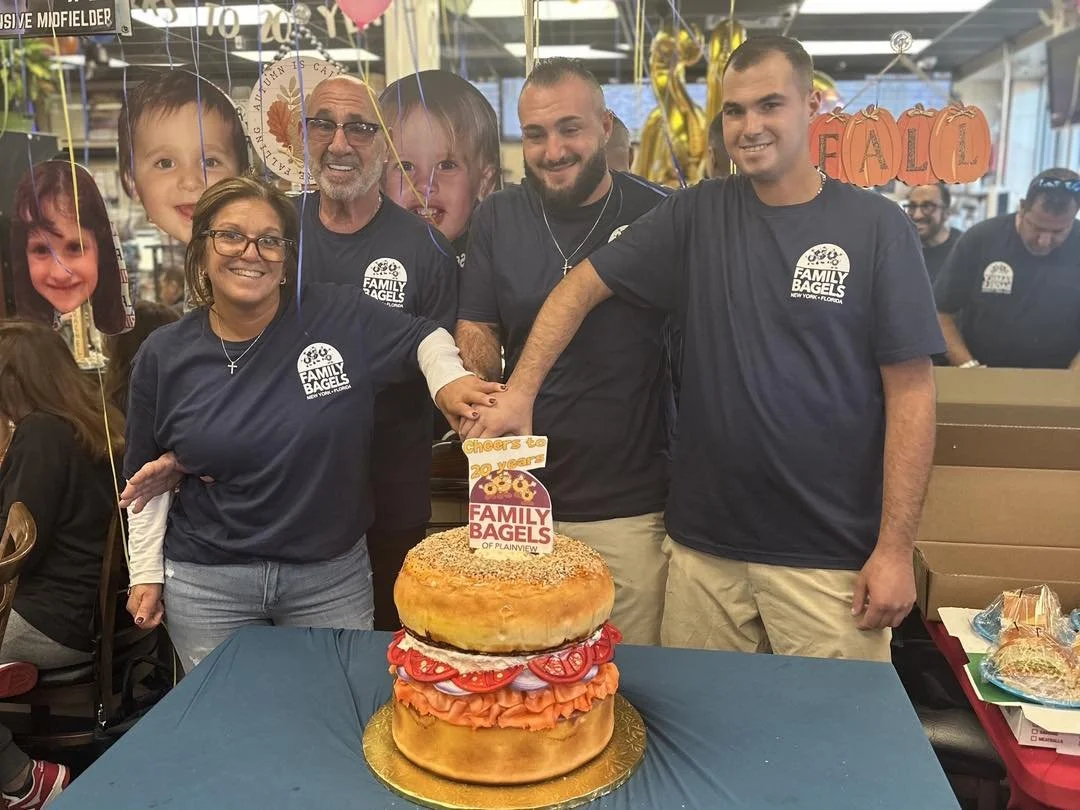 Group of five people celebrating at Family Bagels, standing behind a large bagel cake decorated with strawberries and a sign that says 'Cheers to 20 years'.