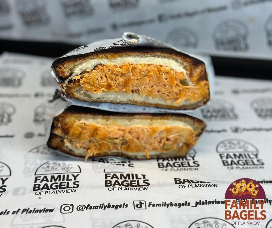 A cross-section of a bagel sandwich with a filling of fried chicken, cream cheese, and pickles, wrapped in branded paper from Family Bagels of Plainview.