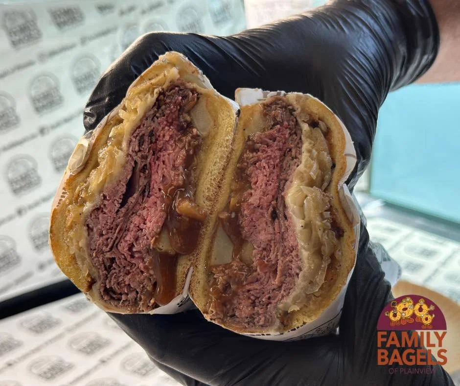 Close-up of a half-cut dip sandwich with layers of cooked ground beef, cheese, onions, and sauce, held by a person wearing a black glove. In the background, there is a logo that says 'Family Bagels of Plainview'.