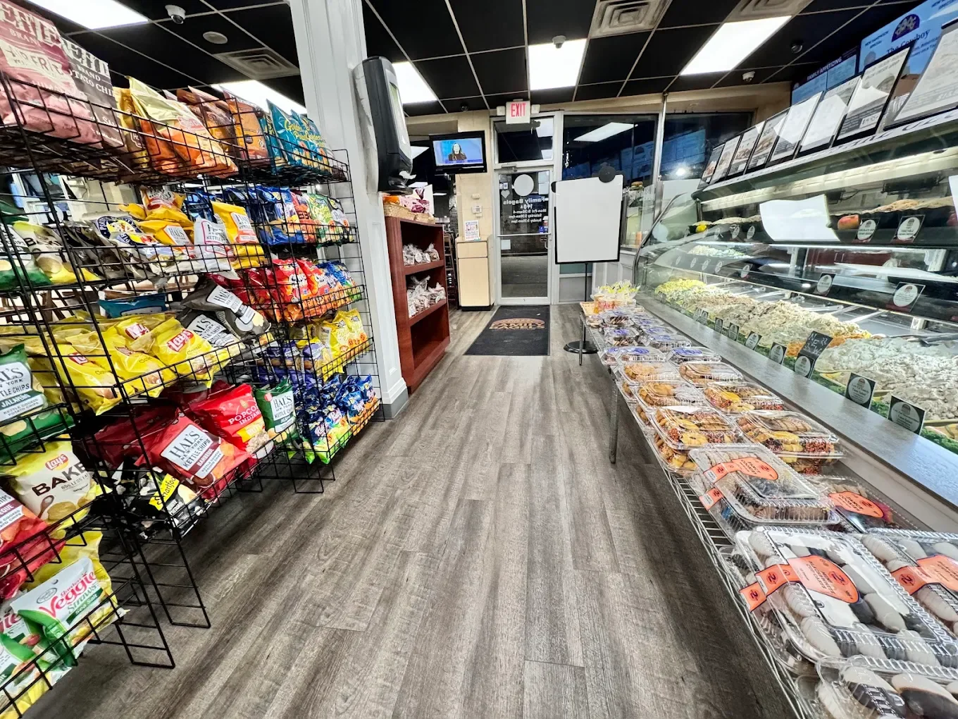 View of a grocery store interior with snack shelves on the left, a display case of prepared foods on the right, and a door at the back.