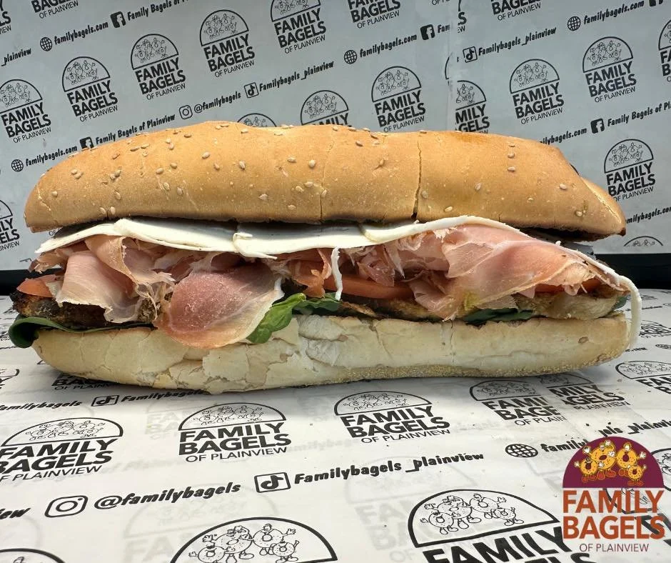 A sandwich with toasted sesame seed bun, containing slices of ham, lettuce, and a fried egg, placed on branded paper from Family Bagels in Plainview.