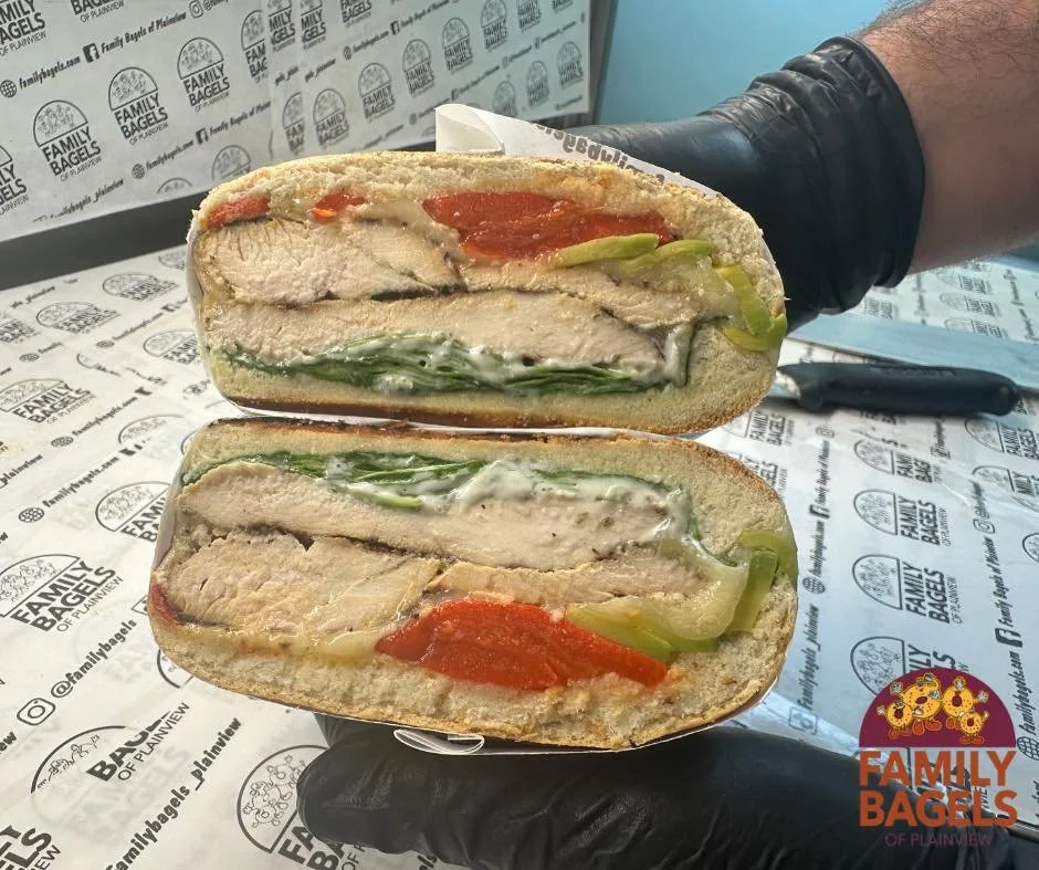 A person holding a halved sandwich with layers of breaded chicken, green bell peppers, and red peppers, inside a toasted bread bun, with a background of patterned paper from Family Bagels.