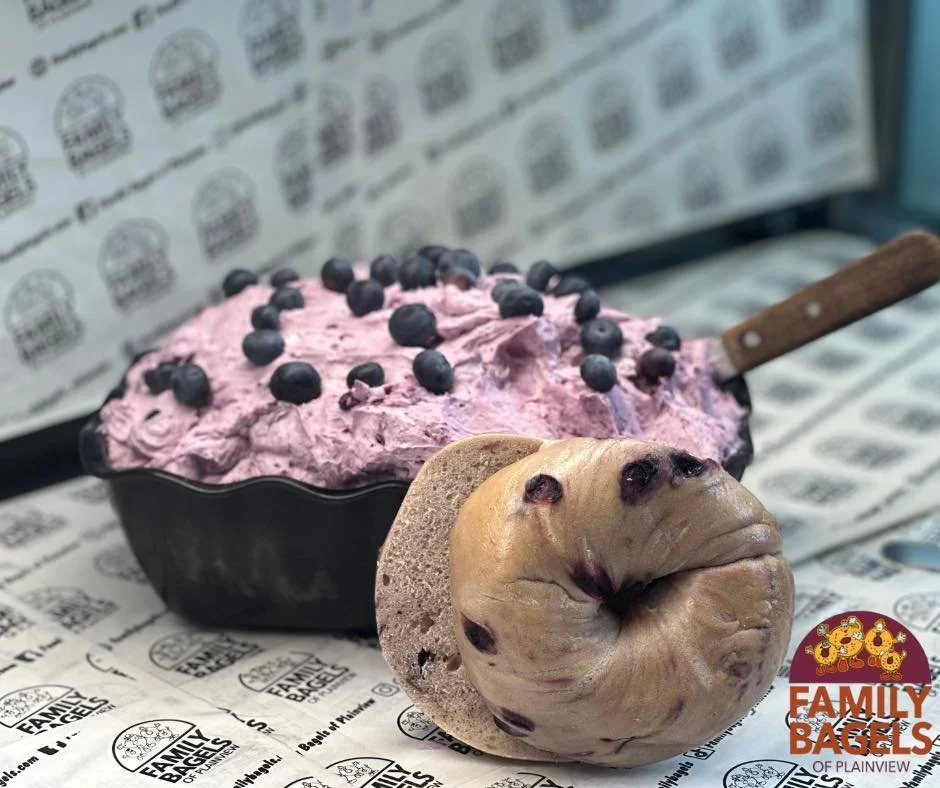 A blueberry ice cream in a black bowl with a wooden spoon, accompanied by a bagel with blueberries in front of it, on branded paper background from Family Bagels.