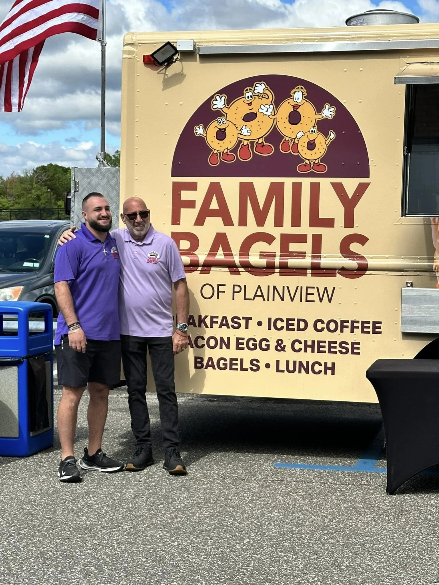 Two men standing in front of a food truck called "Family Bagels of Plainview." The truck features cartoon bagels with arms and legs. The menu lists breakfast, iced coffee, bacon, egg & cheese, bagels, and lunch. One man is wearing a purple shirt and shorts, the other is wearing a light purple shirt and black pants. An American flag is visible in the background.