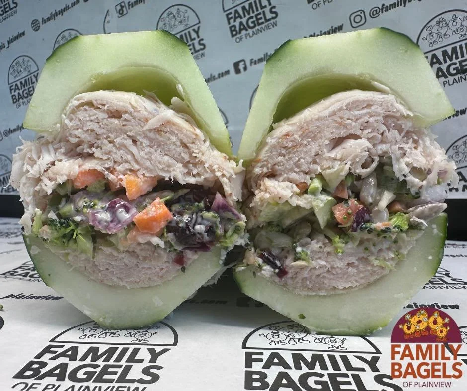 Two halves of a bagel sandwich filled with lettuce, shredded chicken, and vegetables, with cucumber slices on top, on branded paper from Family Bagels.