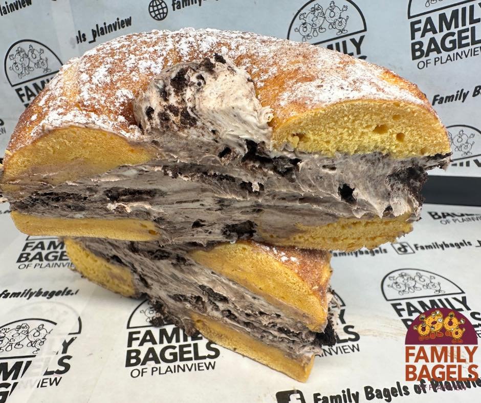 A cross-section of a large, stuffed bagel with cookies and cream flavored cream cheese, visible cookie pieces, and a dusting of powdered sugar, placed on branded paper from Family Bagels of Plainview.