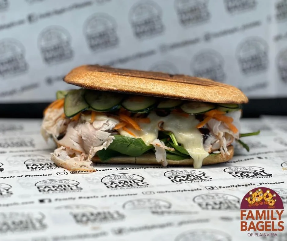 A sandwich with sliced cucumbers, shredded carrots, turkey or chicken, and leafy greens in a toasted bun, served on branded paper with 'Family Bagels' logo.