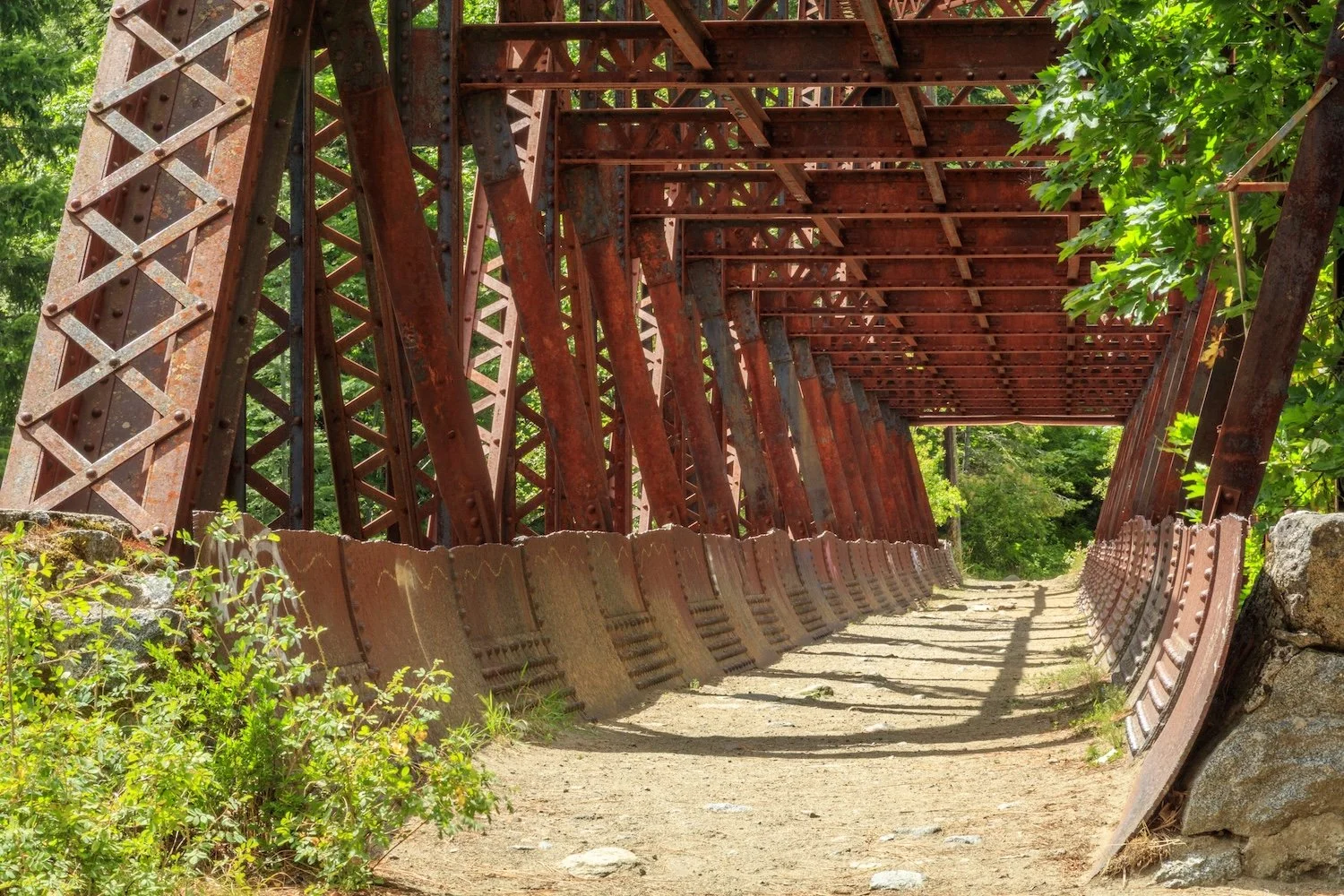 An old, rusted metal bridge with a dirt pathway underneath, surrounded by green trees.
