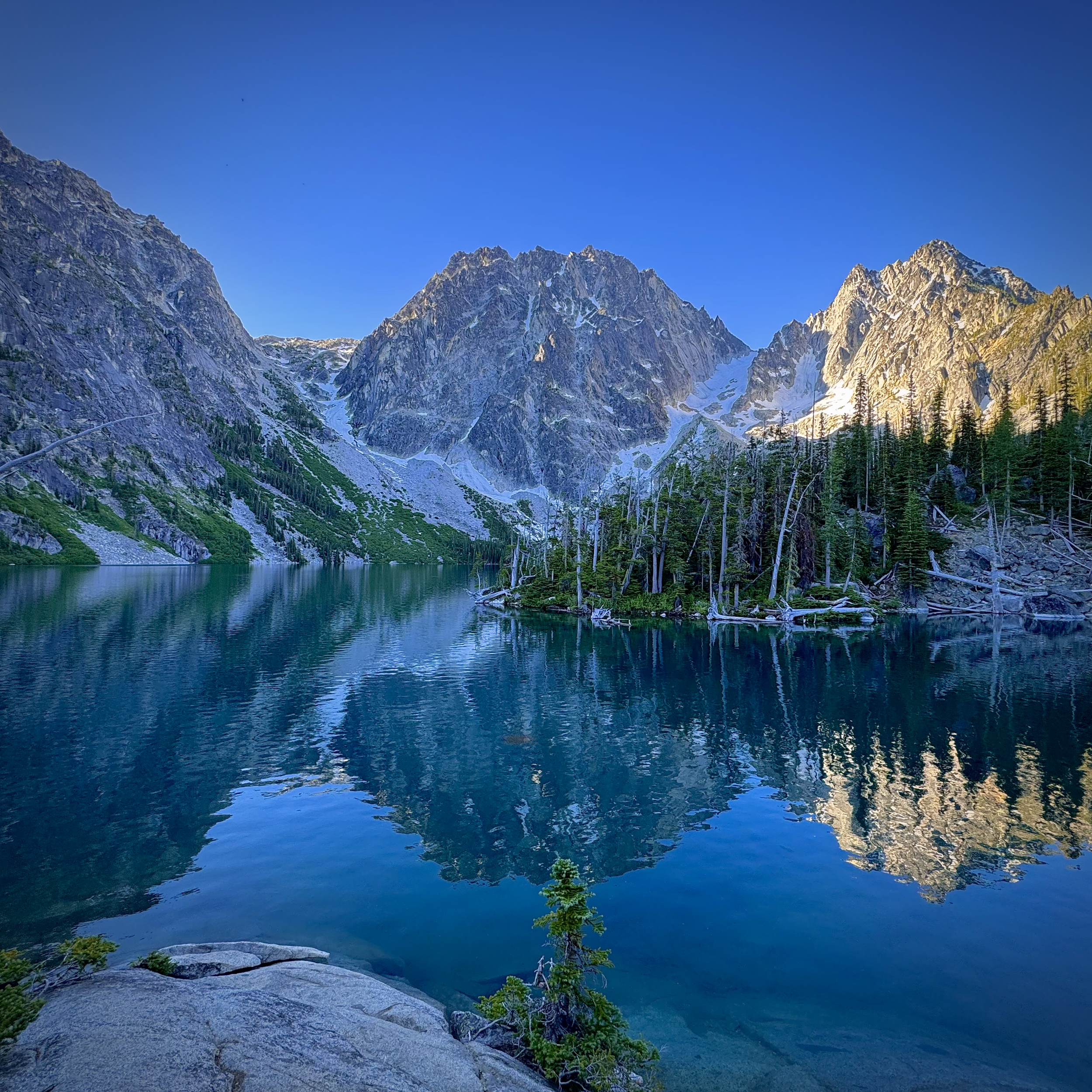 Scenic view of a mountain range with snow patches, a calm lake in the foreground, evergreen trees, and a clear blue sky.